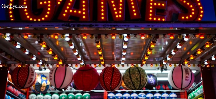 A brightly lit carnival game stall with a large 'GAMES' sign at the top surrounded by red and yellow lights. Various sports balls, including basketballs and soccer balls, are hanging from the ceiling and neatly arranged on shelves below.